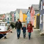 Une famille composée d’un homme, d’une femme, d’un jeune garçon et d’une jeune fille marche sur le port coloré de Saint-Pierre-et-Miquelon, entre maisons en bois et bateaux de pêche, sous une douce lumière de l’Atlantique Nord.