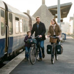 Une famille descend d’un train régional dans une gare normande avec des vélos équipés de sacoches, baignée par la douce lumière du matin.
