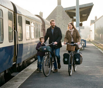 Une famille descend d’un train régional dans une gare normande avec des vélos équipés de sacoches, baignée par la douce lumière du matin.
