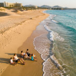 Vue aérienne réaliste d’un homme, d’une femme et de deux enfants sur une plage des Canaries en octobre, avec une eau turquoise, des dunes dorées et un hôtel discret à l’arrière-plan.