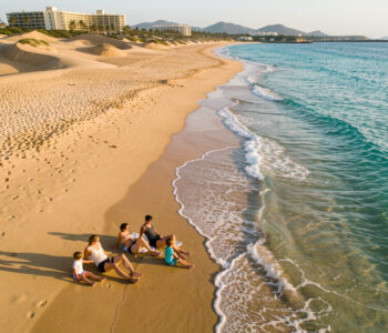 Vue aérienne réaliste d’un homme, d’une femme et de deux enfants sur une plage des Canaries en octobre, avec une eau turquoise, des dunes dorées et un hôtel discret à l’arrière-plan.