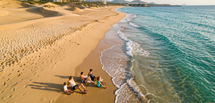 Vue aérienne réaliste d’un homme, d’une femme et de deux enfants sur une plage des Canaries en octobre, avec une eau turquoise, des dunes dorées et un hôtel discret à l’arrière-plan.