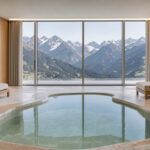 Suite lumineuse d’un spa d’hôtel de luxe avec bassin intérieur en pierre minérale, grandes fenêtres et vue panoramique sur les montagnes alpines.